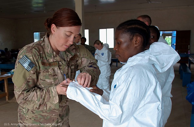 2014年、リベリアの医療従事者がエボラ出血熱の治療エリアに入るため防護スーツ着用を手伝う米空軍のケーシー・ウンターシャー (U.S. Army/Staff Sergeant V. Michelle Woods)